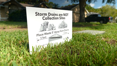 Yard sign placed in the grass near a storm drain with text that says 