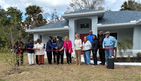 A group of people hold a ceremonial ribbon in front of a new single-family home as two of them hold an oversized pair of scissors to cut it. 