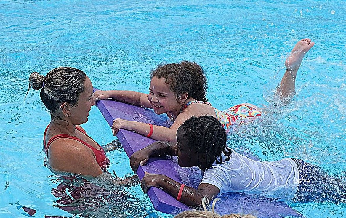 A swim instructor holds a float in a pool while a girl and a boy on the float practice kicking at Eddie Lee Taylor Sr. Pool