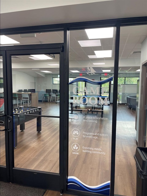 Glass door to newly renovated game room at Joseph N. Davis Community Center. Game room is visible through the doors. Doors have text that says Game Room: A place to socialize and belong. Programs and spaces for every age. Enriching minds, building community.