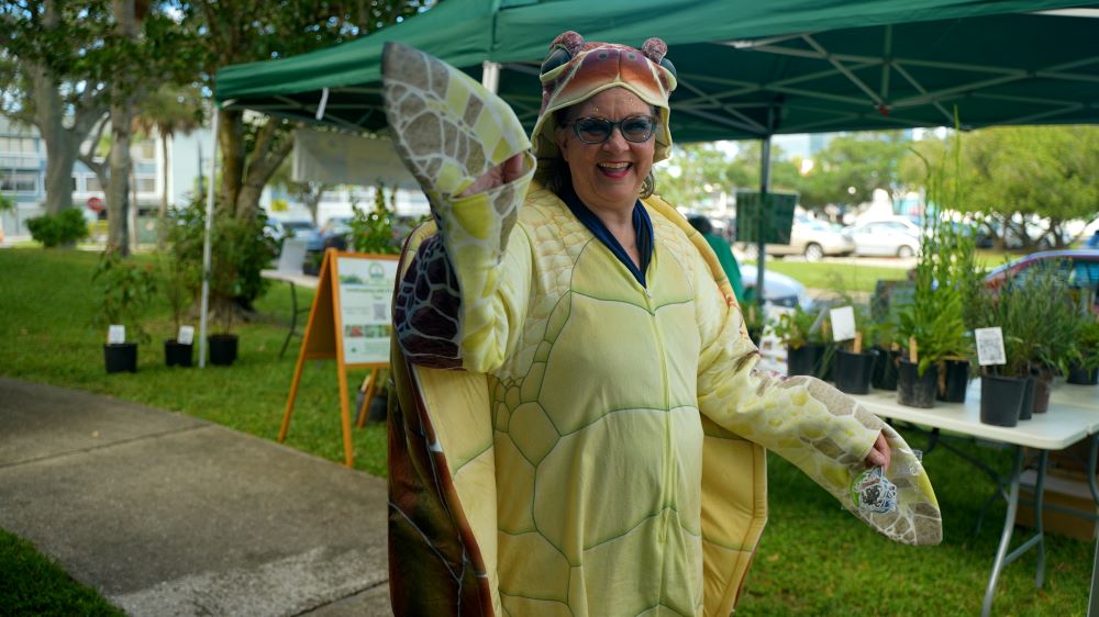 Woman dressed in a sea turtle costume waves in front of native plant exhibit