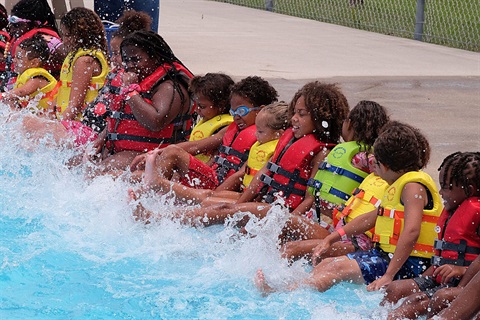 Group of kids wearing life vests and sitting on the edge of a pool splashing with their legs