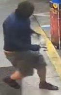 Man in black mask, blue long-sleeved shirt, grey/brown shorts and black shoes walking away from building