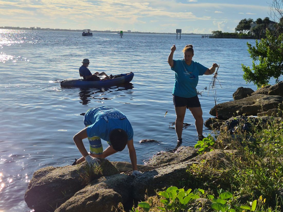 2 volunteers remove litter from rocks at Ballard Park shoreline with kayak in background