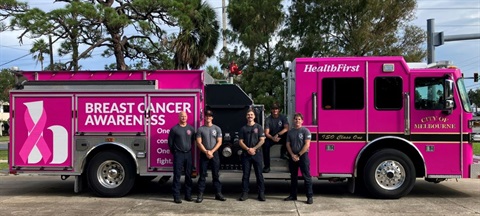 FS 74 Firefighters and Battalion Chief wearing Breast Cancer Awareness Month shirts and standing in front fire engine that has been wrapped in a pink vehicle wrap that says Breast Cancer Awareness and has the Health First logo with a pink ribbon